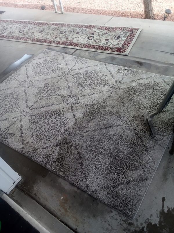 A gray patterned area rug being cleaned with a wand on concrete, one side lighter than the other